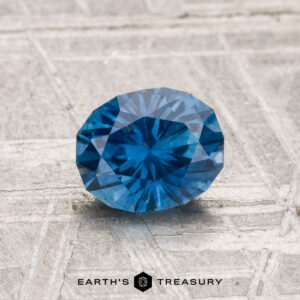 A 1.18-carat heated Montana Sapphire is displayed on a textured light gray surface, with "EARTH'S TREASURY" and a hexagon logo below.