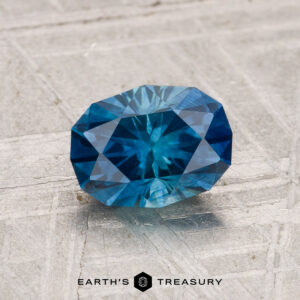 A 1.48-carat heated Montana sapphire with geometric facets rests on a textured gray surface. The "Earth's Treasury" logo is displayed at the bottom.