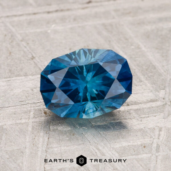 1.48-Carat Montana Sapphire (Heated) A 1.48-carat heated Montana sapphire with geometric facets rests on a textured gray surface. The "Earth's Treasury" logo is displayed at the bottom.
