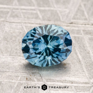 A 1.48-carat heated Montana sapphire is displayed on a textured metallic surface, with the "Earth's Treasury" logo and text at the bottom.