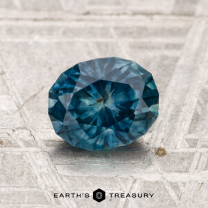 A 1.47-carat, round faceted heated Montana sapphire is displayed on a textured metallic surface with the "EARTH'S TREASURY" logo at the bottom.