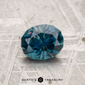 A 1.18-carat faceted Montana sapphire (heated) rests on a textured gray surface, with "EARTH'S TREASURY" displayed below.