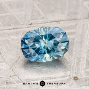 A 1.21-carat heated Montana sapphire with intricate facets is showcased on a textured gray surface, with the Earth's Treasury logo displayed below.