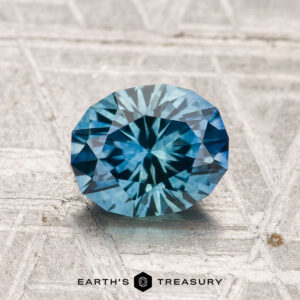 A 1.44-carat heated Montana sapphire with a complex cut rests on a textured gray surface, displaying the "Earth's Treasury" logo and text at the bottom.