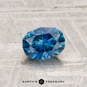A 0.97-carat heated Montana sapphire with blue facets rests on a light textured surface, with the Earth's Treasury logo and text shown below.