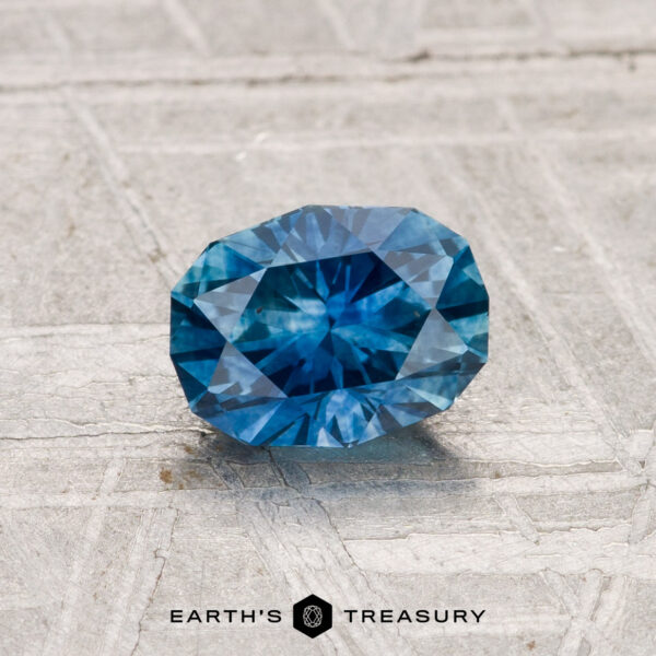 0.97-Carat Montana Sapphire (Heated) A 0.97-carat heated Montana sapphire with blue facets rests on a light textured surface, with the Earth's Treasury logo and text shown below.