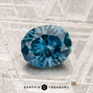 A 1.41-carat heated Montana sapphire, faceted in an oval blue-green shape, is shown on a textured gray surface with "EARTH'S TREASURY" at the bottom.