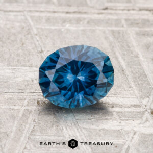 A 1.17-carat heated Montana Sapphire, faceted in an oval cut with a vivid blue hue, is displayed on a textured gray surface with "Earth's Treasury" at the bottom.