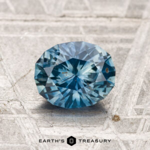 A 1.41-carat heated Montana sapphire, cut in a faceted oval shape, is displayed on a textured gray surface with "EARTH'S TREASURY" at the bottom.
