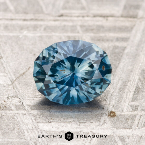 1.41-Carat Montana Sapphire (Heated) A 1.41-carat heated Montana sapphire, cut in a faceted oval shape, is displayed on a textured gray surface with "EARTH'S TREASURY" at the bottom.