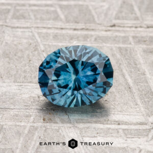 A 1.16-carat heated Montana Sapphire, round brilliant-cut and blue, is displayed on a textured gray surface with "EARTH'S TREASURY" at the bottom.