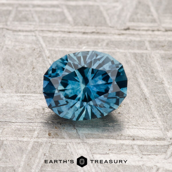 1.16-Carat Montana Sapphire (Heated) A 1.16-carat heated Montana Sapphire, round brilliant-cut and blue, is displayed on a textured gray surface with "EARTH'S TREASURY" at the bottom.