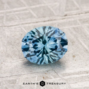 A 1.26-carat heated Montana Sapphire, round brilliant-cut and blue, displayed on a textured light gray surface with the "Earth's Treasury" logo and text at the bottom.