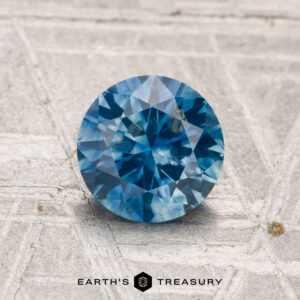 1.30-carat heated Montana sapphire in round brilliant cut shown on a textured light gray surface, with "EARTH'S TREASURY" displayed at the bottom.