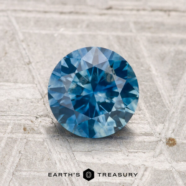 1.30-Carat Montana Sapphire (Heated) 1.30-carat heated Montana sapphire in round brilliant cut shown on a textured light gray surface, with "EARTH'S TREASURY" displayed at the bottom.