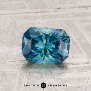 1.46-carat heated Montana sapphire with a complex faceted cut, rectangular shape, and blue hue, displayed on a light textured surface with "EARTH'S TREASURY" below.