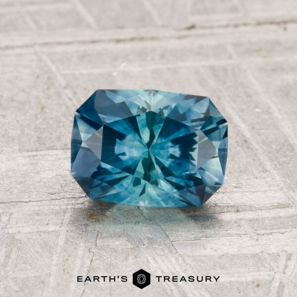 1.46-Carat Montana Sapphire (Heated) 1.46-carat heated Montana sapphire with a complex faceted cut, rectangular shape, and blue hue, displayed on a light textured surface with "EARTH'S TREASURY" below.