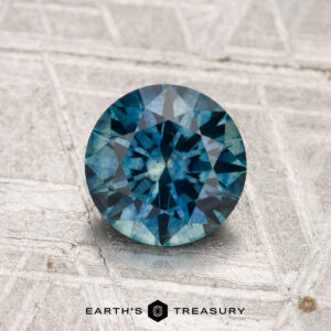 A 1.27-carat heated Montana sapphire, round and faceted, sits on a textured metallic surface with "EARTH'S TREASURY" beneath.