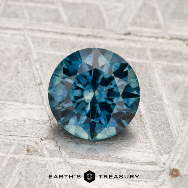 1.27-Carat Montana Sapphire (Heated) A 1.27-carat heated Montana sapphire, round and faceted, sits on a textured metallic surface with "EARTH'S TREASURY" beneath.