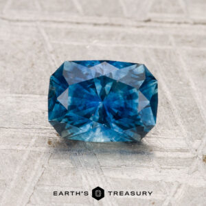 A 1.42-carat heated Montana sapphire with multiple facets is shown on a light grey textured surface. The text "EARTH'S TREASURY" appears at the bottom of the image.