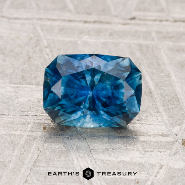 1.42-Carat Montana Sapphire (Heated) A 1.42-carat heated Montana sapphire with multiple facets is shown on a light grey textured surface. The text "EARTH'S TREASURY" appears at the bottom of the image.