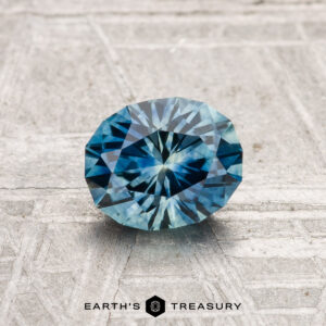 A 1.13-carat heated Montana Sapphire, round and faceted, is shown on a textured light gray surface with the Earth's Treasury logo at the bottom.