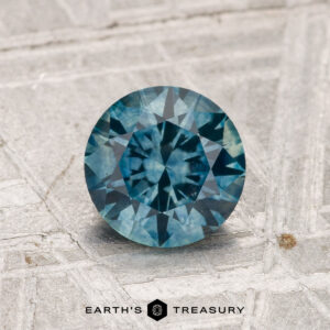 1.25-Carat Montana Sapphire (Heated), round brilliant-cut, displayed on a textured light gray surface with the “Earth’s Treasury” logo at the bottom.