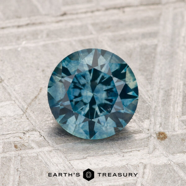 1.25-Carat Montana Sapphire (Heated) 1.25-Carat Montana Sapphire (Heated), round brilliant-cut, displayed on a textured light gray surface with the “Earth’s Treasury” logo at the bottom.