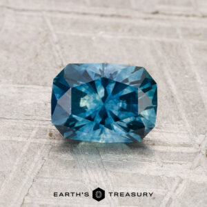 A 1.21-carat heated Montana sapphire, cut in a rectangular blue-green facet, is displayed on a gray textured surface featuring the "Earth's Treasury" logo at the bottom.