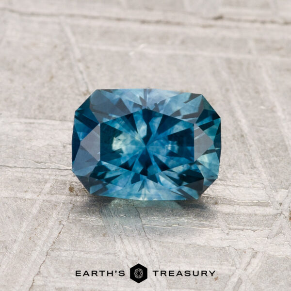 1.21-Carat Montana Sapphire (Heated) A 1.21-carat heated Montana sapphire, cut in a rectangular blue-green facet, is displayed on a gray textured surface featuring the "Earth's Treasury" logo at the bottom.