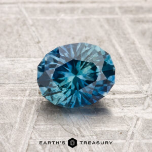 A 1.35-carat heated Montana sapphire, round and faceted, is shown on a textured gray surface with "Earth's Treasury" beneath it.