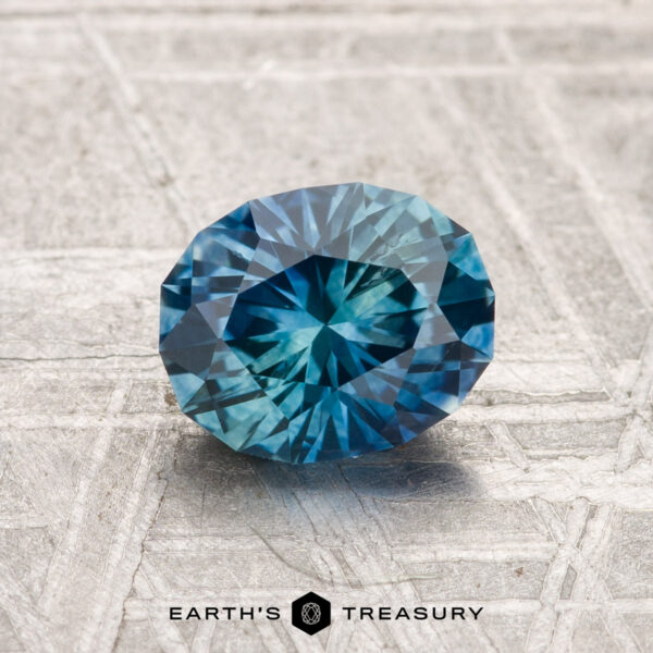 1.35-Carat Montana Sapphire (Heated) A 1.35-carat heated Montana sapphire, round and faceted, is shown on a textured gray surface with "Earth's Treasury" beneath it.