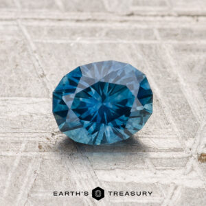 A round, faceted 1.08-carat heated Montana sapphire with a vivid blue hue rests on a textured light surface, the "Earth's Treasury" logo displayed below.