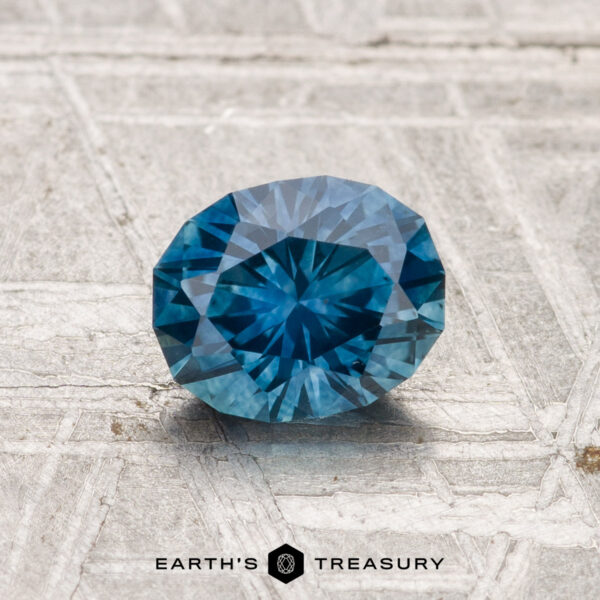 1.08-Carat Montana Sapphire (Heated) A round, faceted 1.08-carat heated Montana sapphire with a vivid blue hue rests on a textured light surface, the "Earth's Treasury" logo displayed below.