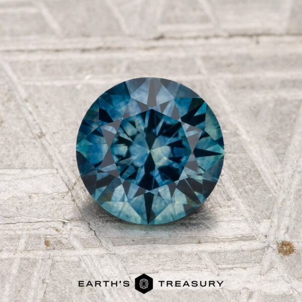 1.22-Carat Montana Sapphire (Heated) A 1.22-carat heated Montana sapphire in round brilliant cut is displayed on a textured gray surface with the "Earth's Treasury" logo below.