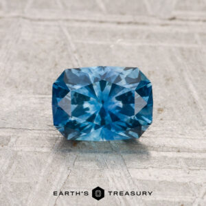 A rectangular, faceted blue 1.07-carat heated Montana sapphire rests on a textured light surface with "EARTH’S TREASURY" below it.