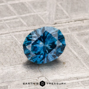 A 1.08-carat round-cut heated Montana sapphire is displayed on a textured grey surface, featuring the "Earth's Treasury" logo at the bottom.