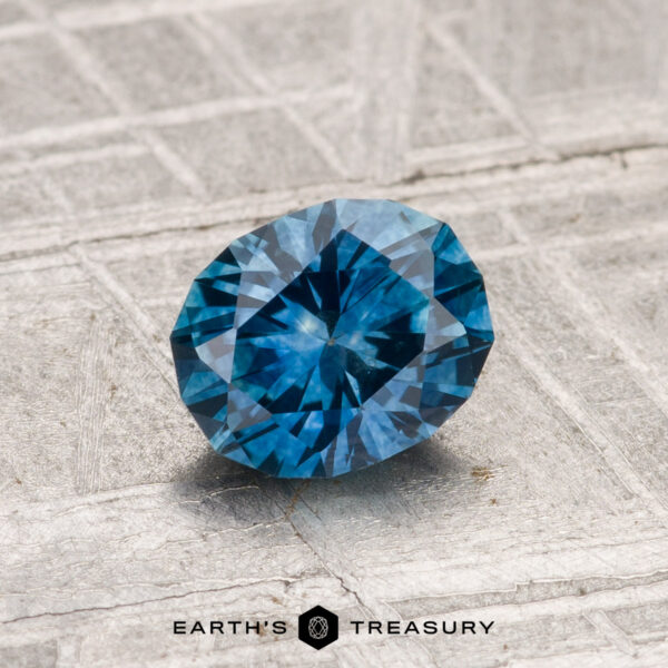 1.08-Carat Montana Sapphire (Heated) A 1.08-carat round-cut heated Montana sapphire is displayed on a textured grey surface, featuring the "Earth's Treasury" logo at the bottom.