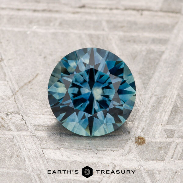 1.21-Carat Montana Sapphire (Heated) A round, faceted blue-green 1.21-carat Montana sapphire (heated) is displayed on a textured pale gray surface with "EARTH'S TREASURY" text and logo at the bottom.