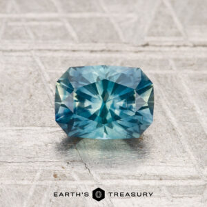 A 1.05-carat heated Montana sapphire with a radiant cut and blue-green hue is displayed on a textured light gray surface, with "EARTH'S TREASURY" below.