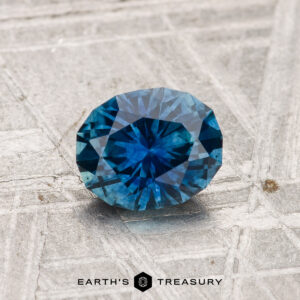 A 1.07-carat heated Montana sapphire, round-cut and blue, rests on a textured metal surface featuring "EARTH'S TREASURY" and a hexagon logo at the bottom of the image.