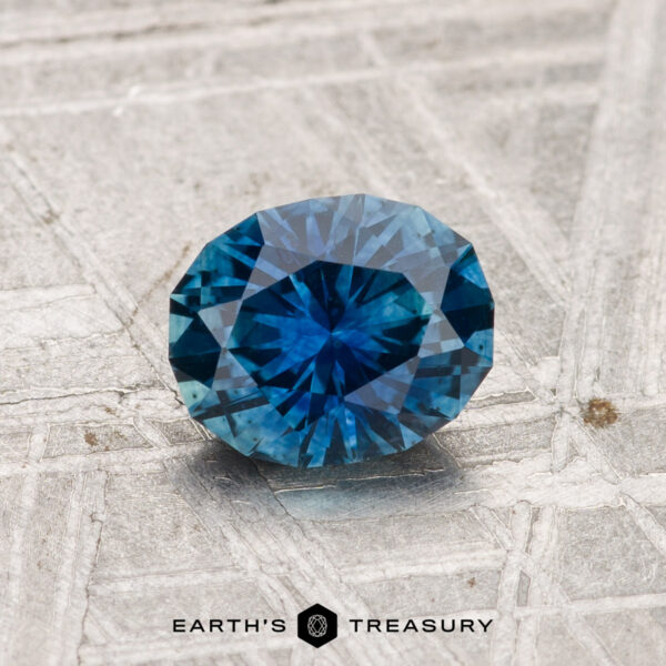 1.07-Carat Montana Sapphire (Heated) A 1.07-carat heated Montana sapphire, round-cut and blue, rests on a textured metal surface featuring "EARTH'S TREASURY" and a hexagon logo at the bottom of the image.