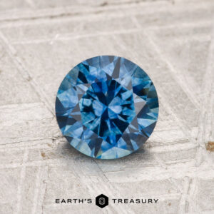 A 1.21-carat heated Montana sapphire in round brilliant cut, shown on a textured light gray surface with the "Earth's Treasury" logo and text below.
