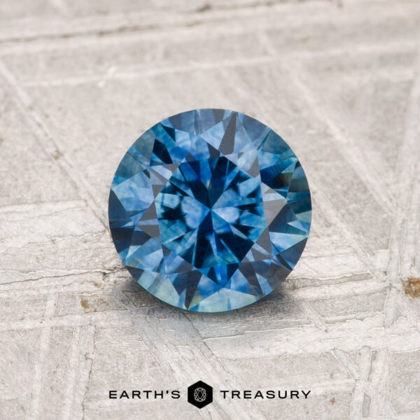 1.21-Carat Montana Sapphire (Heated) A 1.21-carat heated Montana sapphire in round brilliant cut, shown on a textured light gray surface with the "Earth's Treasury" logo and text below.