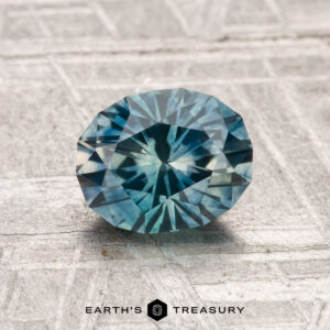 A 1.35-carat heated Montana sapphire, round and faceted in a blue-green hue, is displayed on a textured light gray surface with the Earth’s Treasury logo at the bottom.