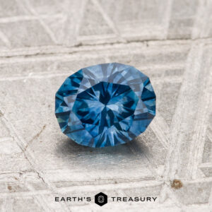 A 1.03-carat heated Montana sapphire with a round brilliant cut rests on a textured metallic surface featuring the “Earth’s Treasury” logo at the bottom.