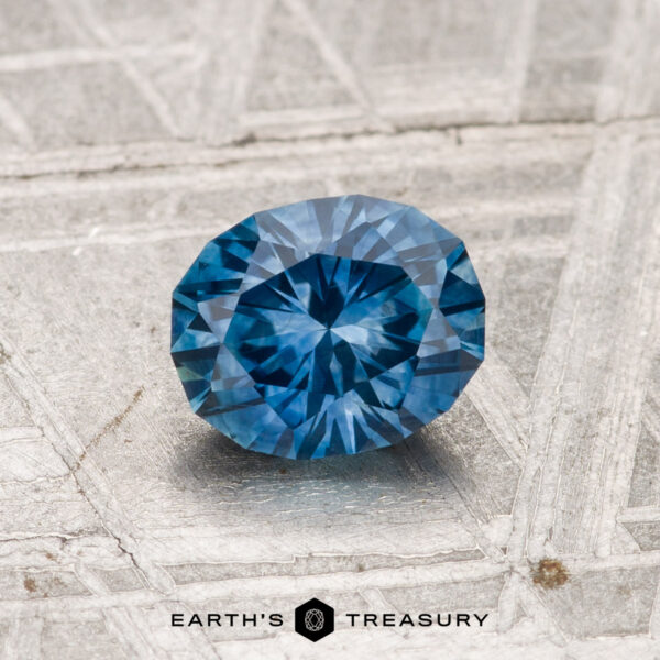 1.03-Carat Montana Sapphire (Heated) A 1.03-carat heated Montana sapphire with a round brilliant cut rests on a textured metallic surface featuring the “Earth’s Treasury” logo at the bottom.
