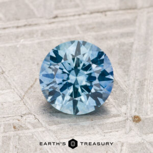 A 1.19-carat heated Montana Sapphire with a round brilliant cut is displayed on a textured gray surface, with "EARTH'S TREASURY" shown at the bottom.