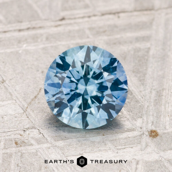 1.19-Carat Montana Sapphire (Heated) A 1.19-carat heated Montana Sapphire with a round brilliant cut is displayed on a textured gray surface, with "EARTH'S TREASURY" shown at the bottom.