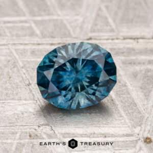 A 1.34-carat heated Montana sapphire, faceted in an oval shape, rests on a textured metallic surface with "EARTH'S TREASURY" and a logo visible at the bottom.